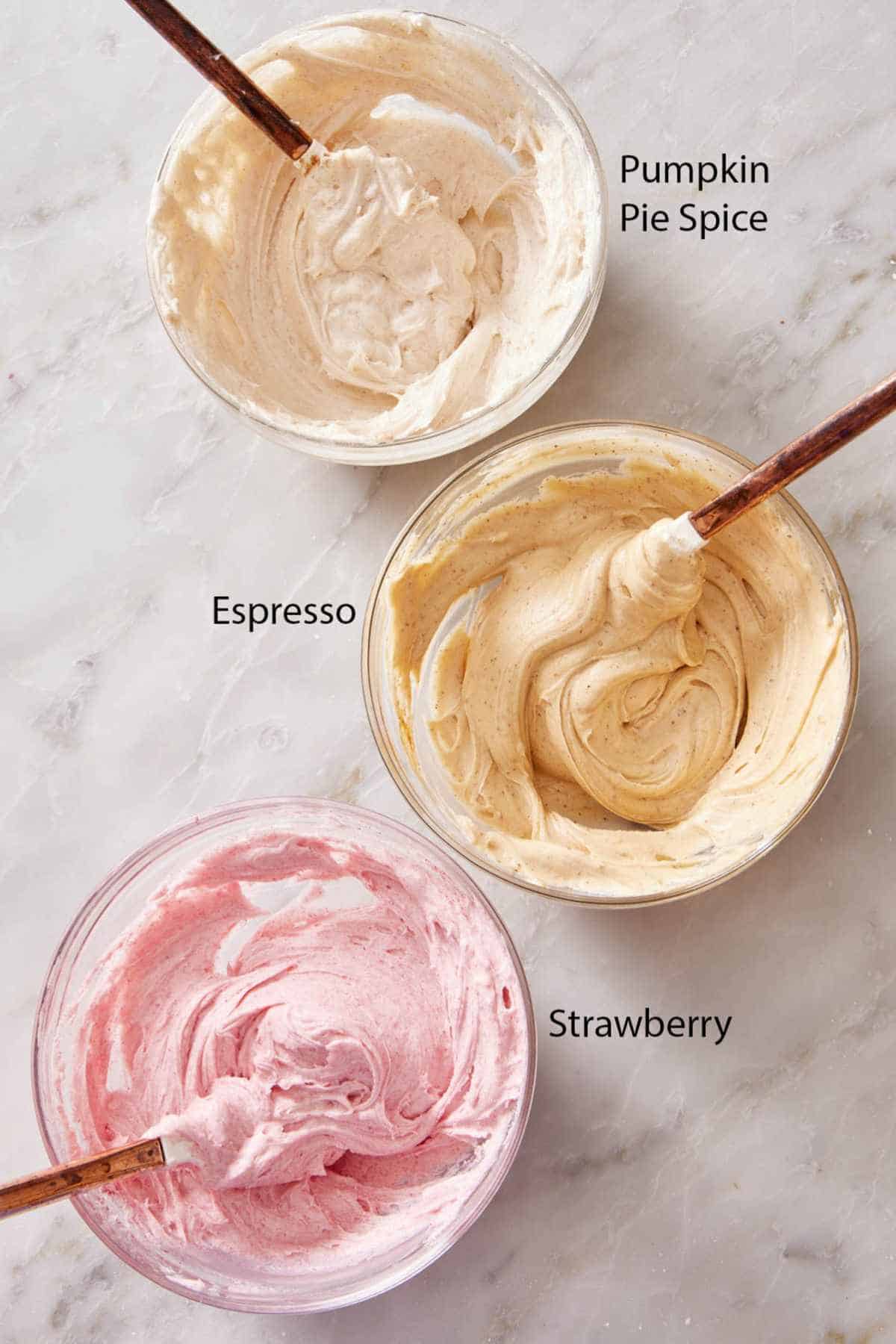 Overhead shot of 3 bowls containing cream cheese frosting flavor variations: pumpkin pie spice, espresso, and strawberry.