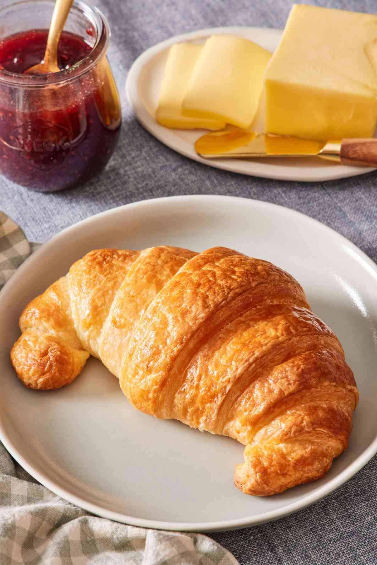 A buttery croissant on a plate with butter and jam in the background.