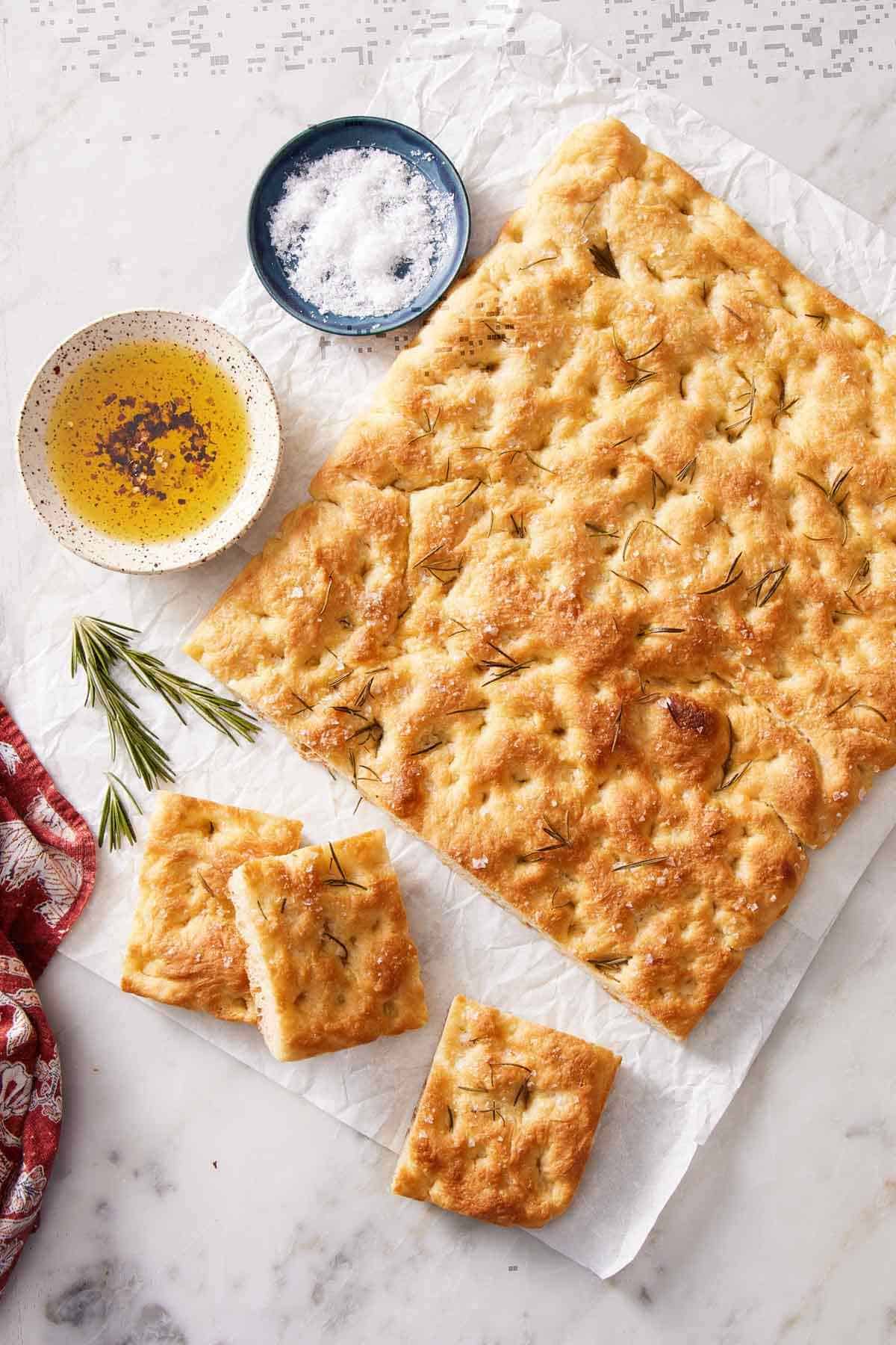 Overhead shot of focaccia bread with a few slices cut out. Dipping oil, salt, and rosemary sprigs next to it.