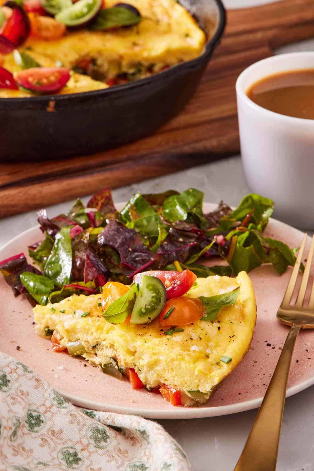 A slice of frittata on a plate with a leafy green salad on the side. A cup of coffee and the rest of the frittata also in frame.