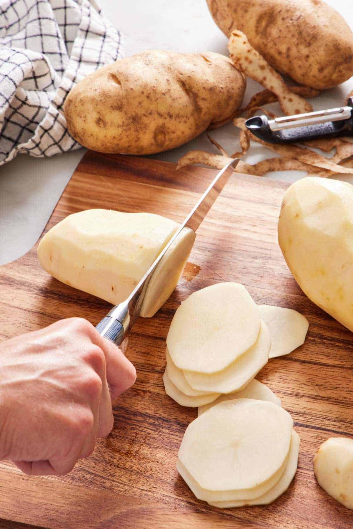 Potatoes sliced on a cutting board with more whole potatoes off to the side.