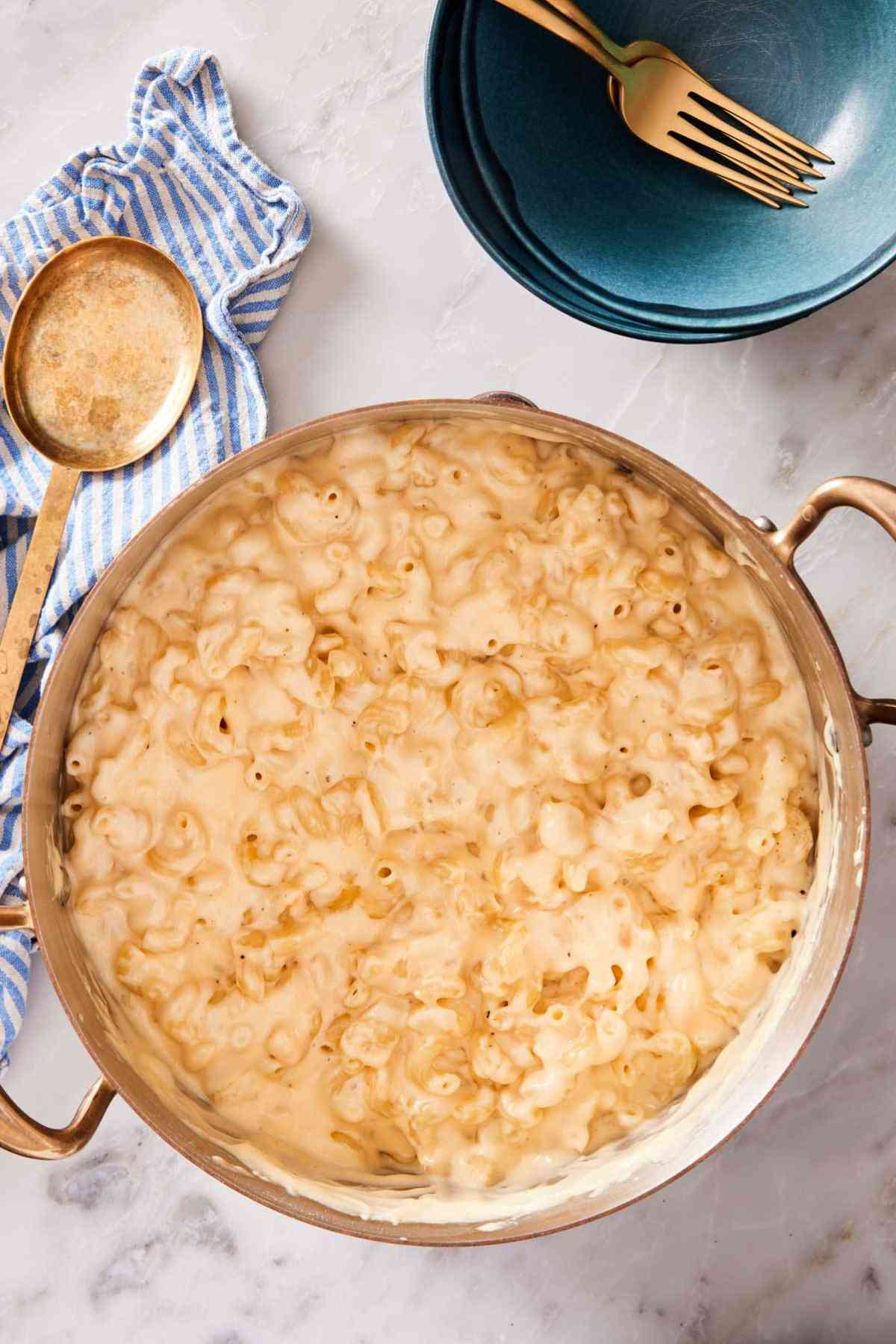 Overhead shot of mac and cheese in a large pot on a marble countertop with flatware and a striped table linen around it.