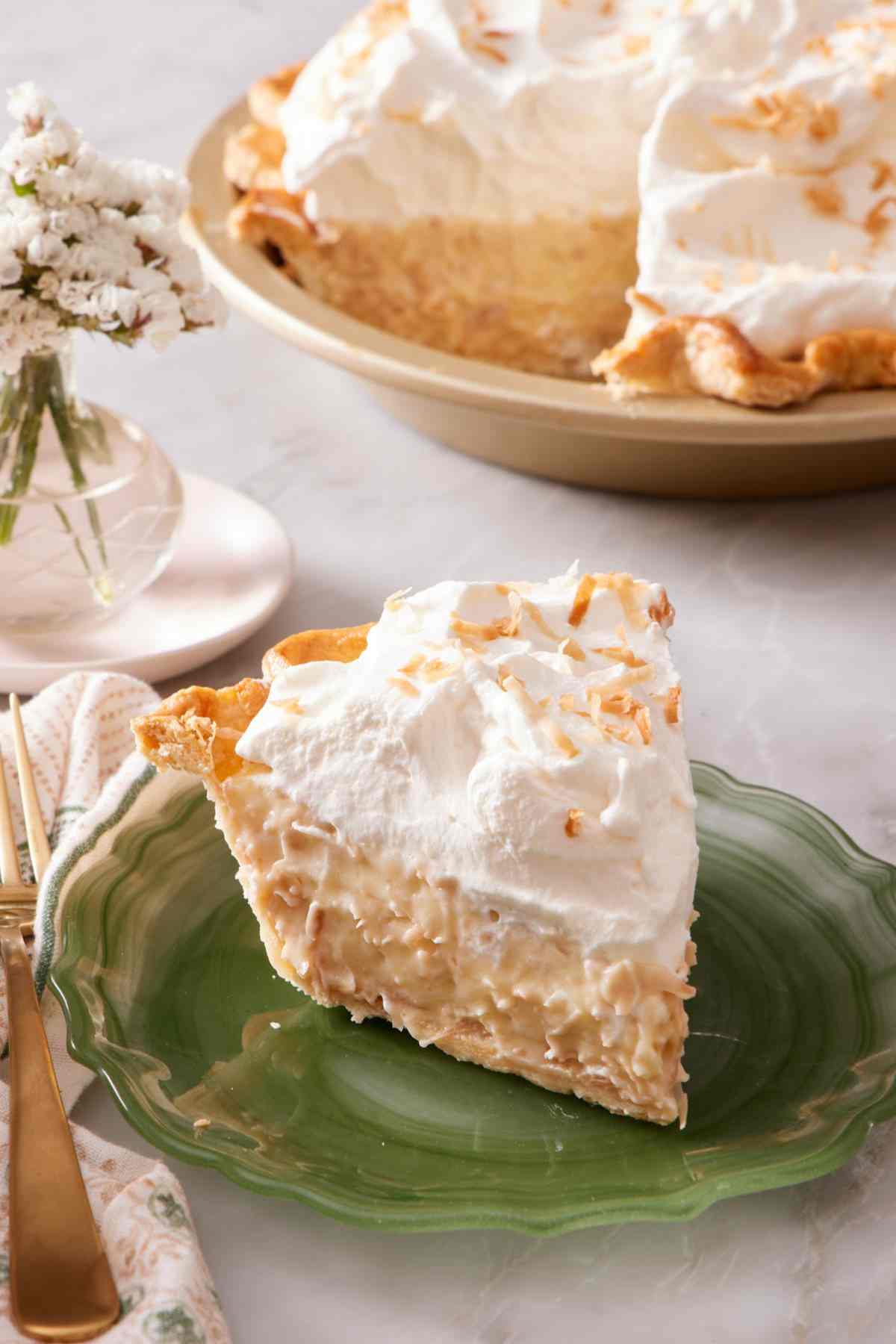 A slice of homemade coconut cream pie on a green plate with the rest of the pie in the background.