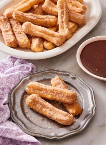 Air fryer churros on a plate, with more on a platter behind it, as well as a bowl of chocolate dipping sauce.