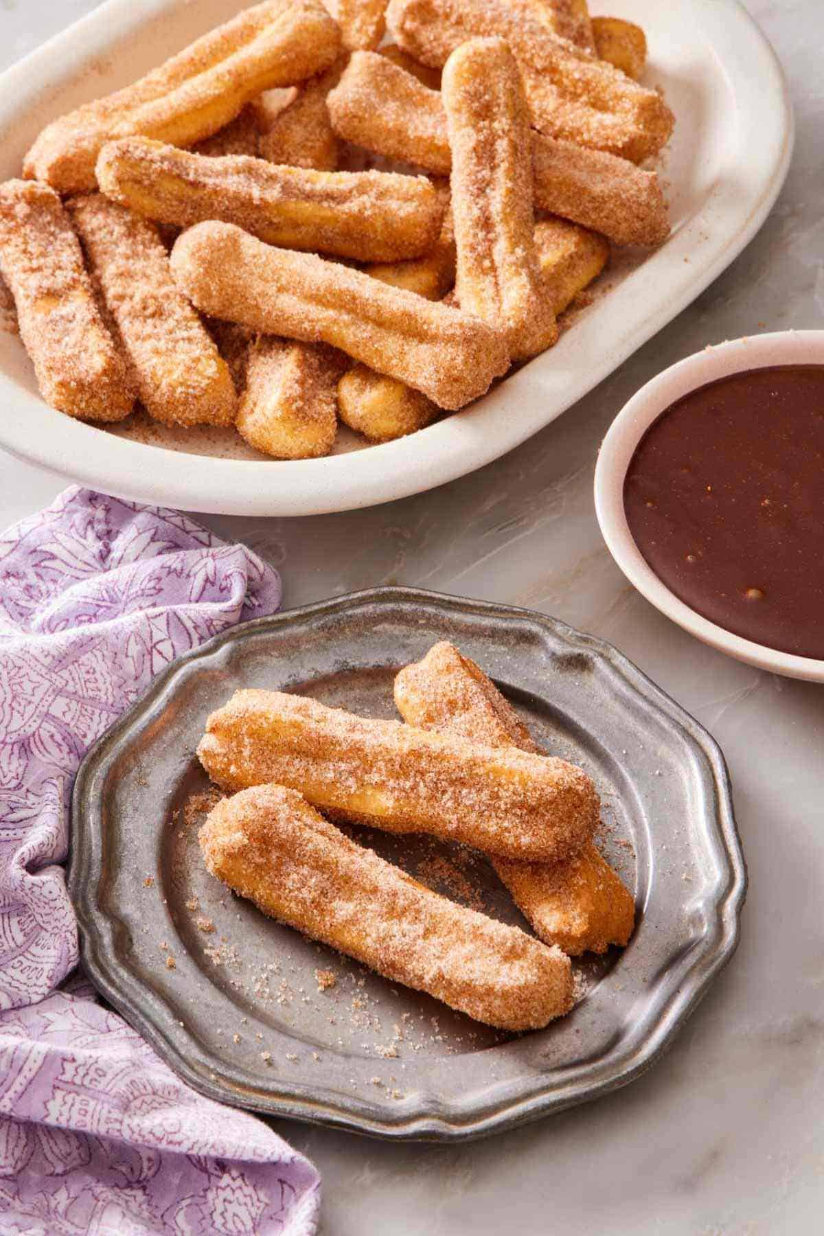 Air fryer churros on a plate, with more on a platter behind it, as well as a bowl of chocolate dipping sauce.