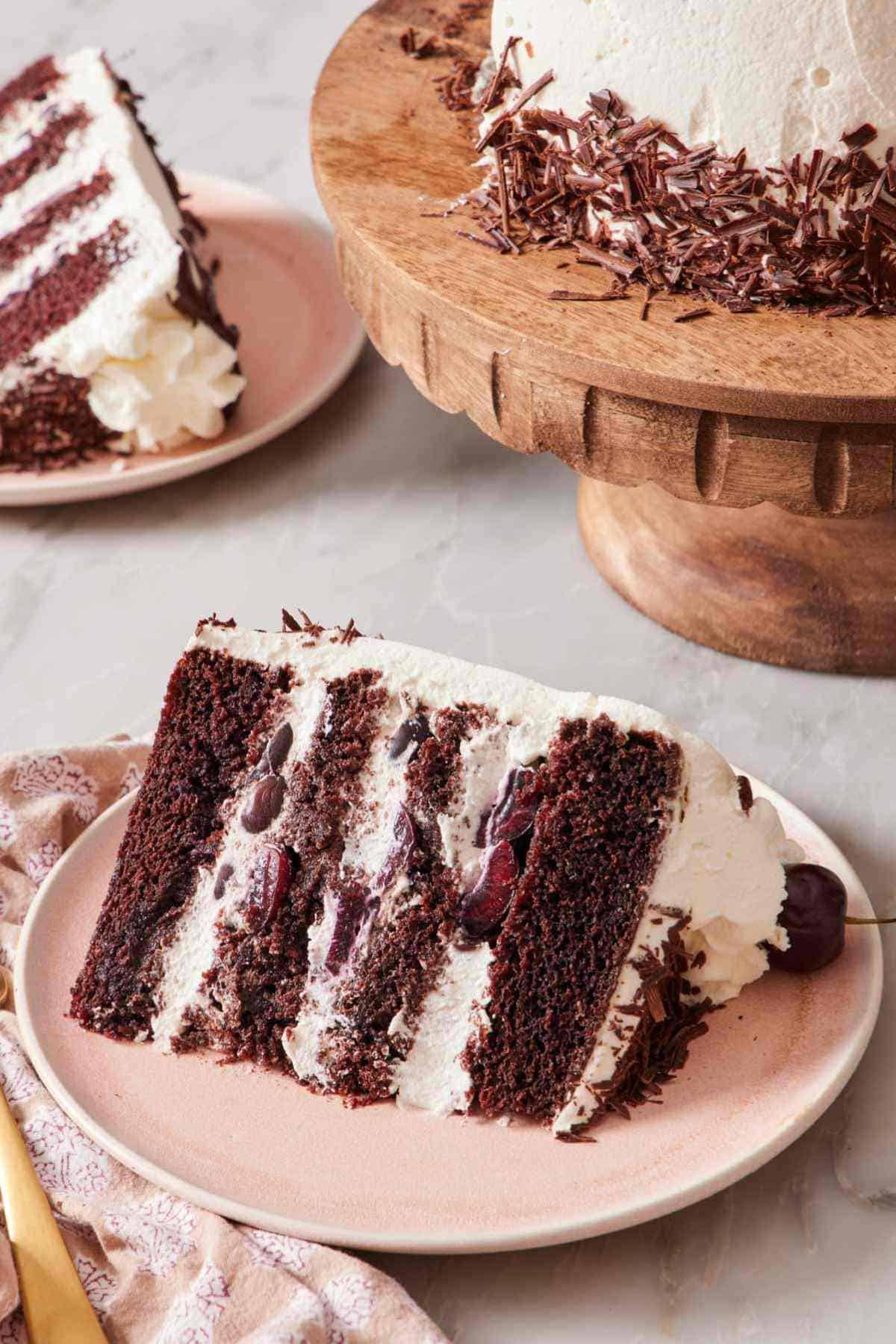 A slice of black forest cake on a plate, with another slice and the rest of the cake mostly out of frame in the background.