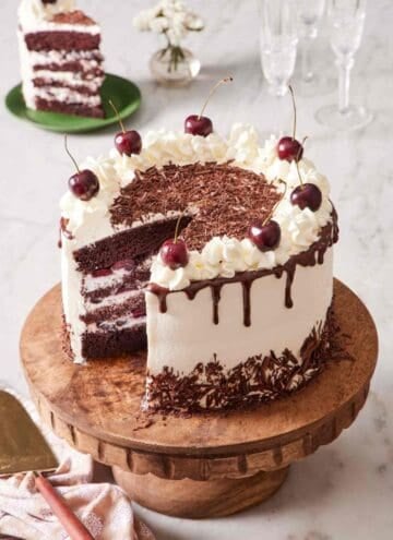 A black forest cake on a wooden cake stand with a slice cue out. The slice is on a small serving plate in the background.