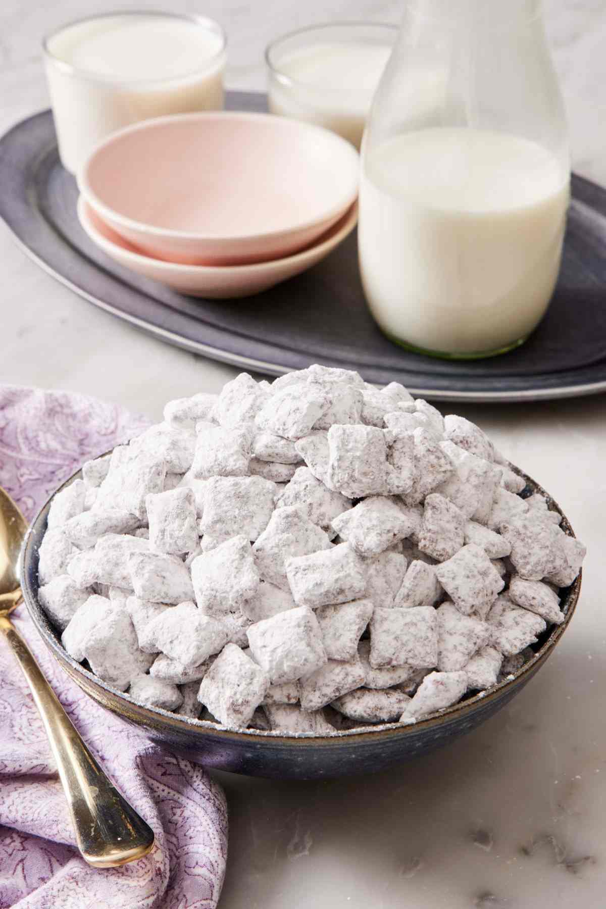 A bowl piled high with homemade puppy chow and a jug of milk with glasses and small bowls in the background.
