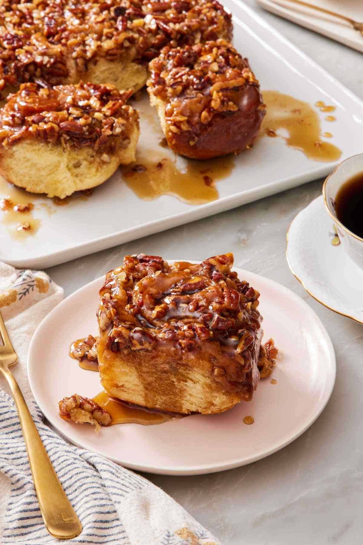 A pecan sticky bun on a white plate with the rest of the sticky buns mostly out of frame.