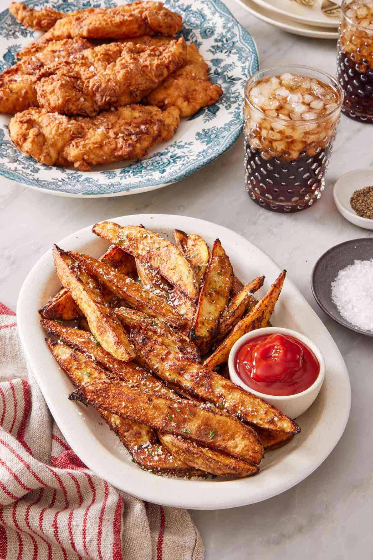 Baked potato wedges on a serving platter with a small dish of ketchup, with fried chicken and two glasses of soda.