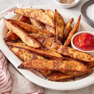 Oven baked potato wedges on a white platter with ketchup. Salt and pepper in the background.