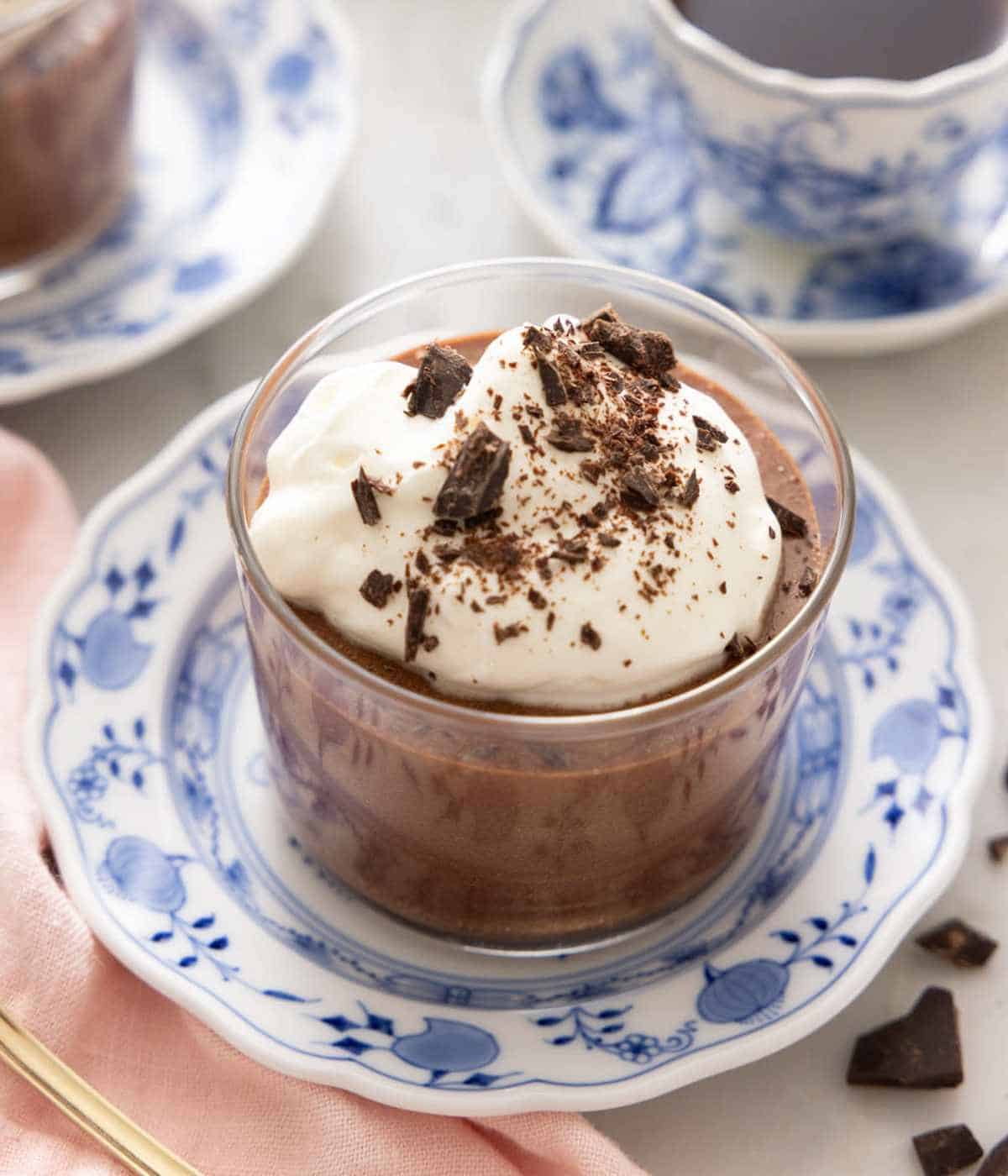 A small glass of chocolate mousse topped with whipped cream and chocolate shavings, set on a blue and white plate.