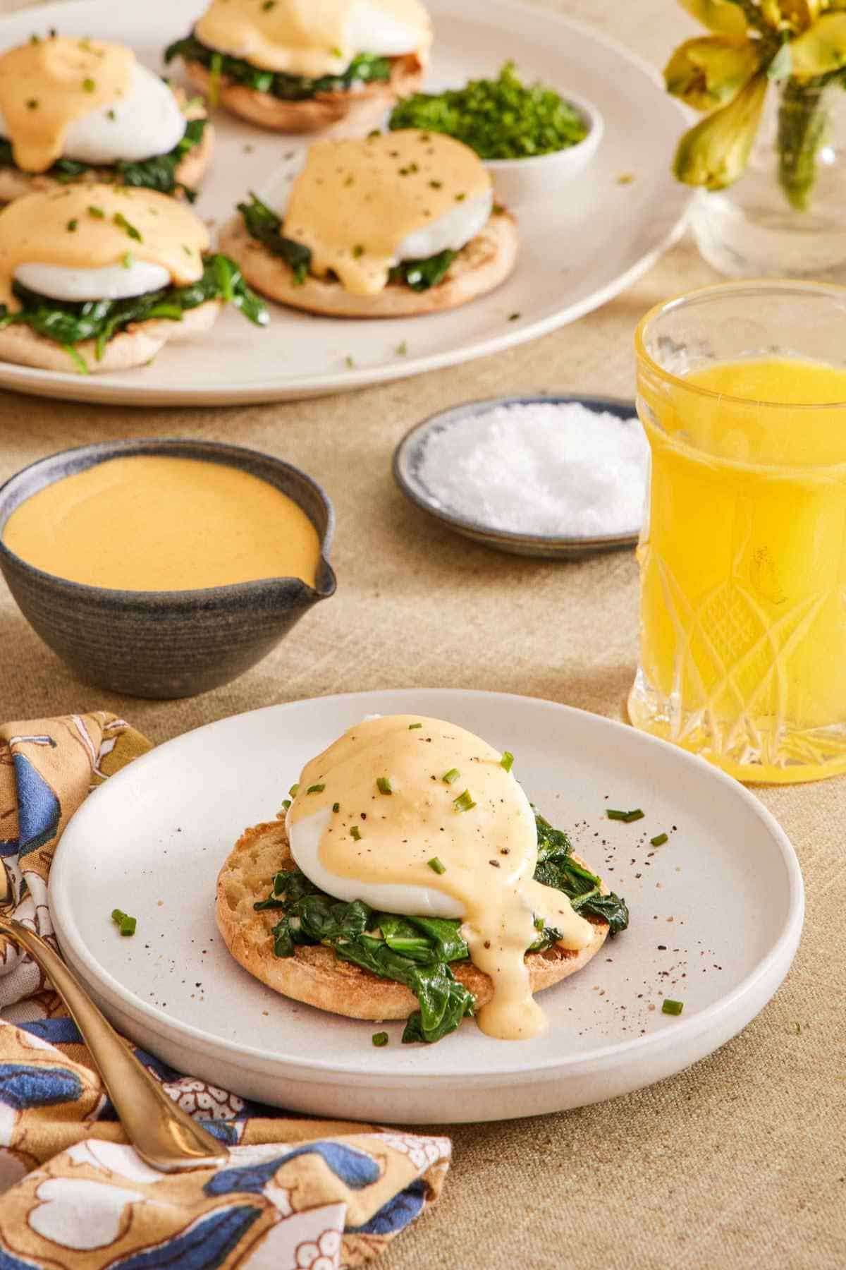Eggs Florentine on a white plate next to a glass of juice, with hollandaise sauce, salt, and more eggs Florentine in the background.