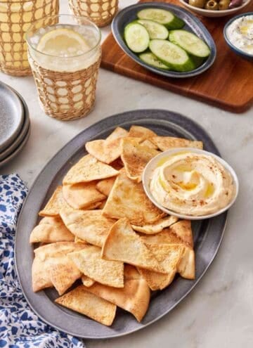Homemade pita chips on a platter with hummus, with sliced cucumber, olives, and tzatziki in the background. A few drinks are next to the platter.