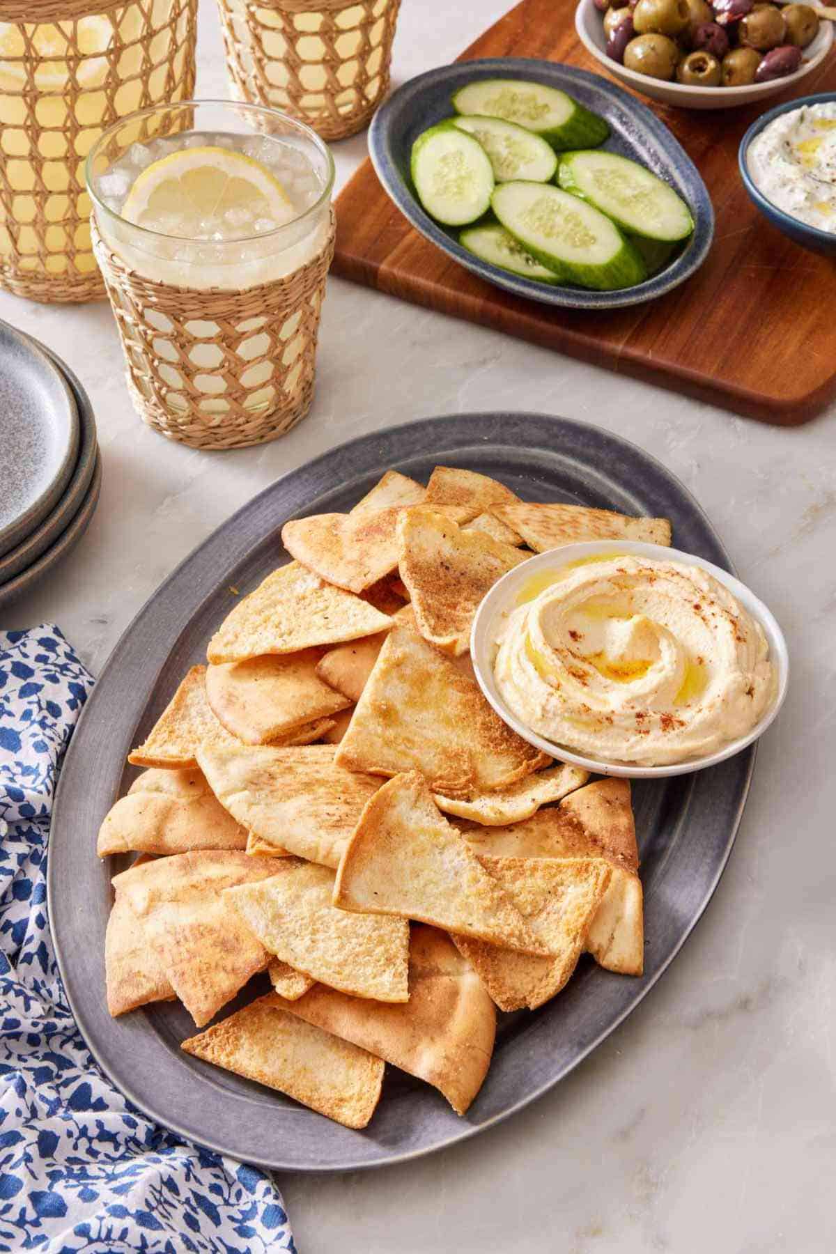 Homemade pita chips on a platter with hummus, with sliced cucumber, olives, and tzatziki in the background. A few drinks are next to the platter.