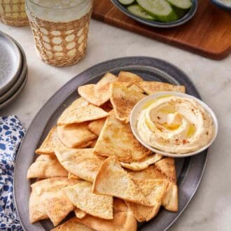 Pinterest graphic showing a large serving platter of homemade pita chips with hummus, with sliced cucumber and drinks in the background.