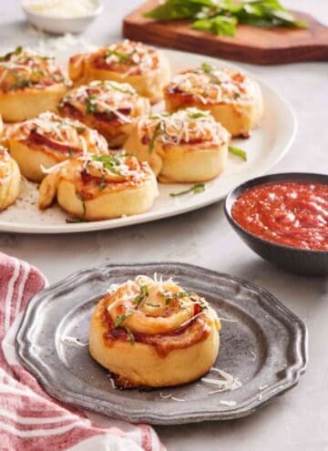 A pizza roll on a grey plate with a bowl of sauce to the side, with more homemade pizza rolls slightly out of focus in the background.