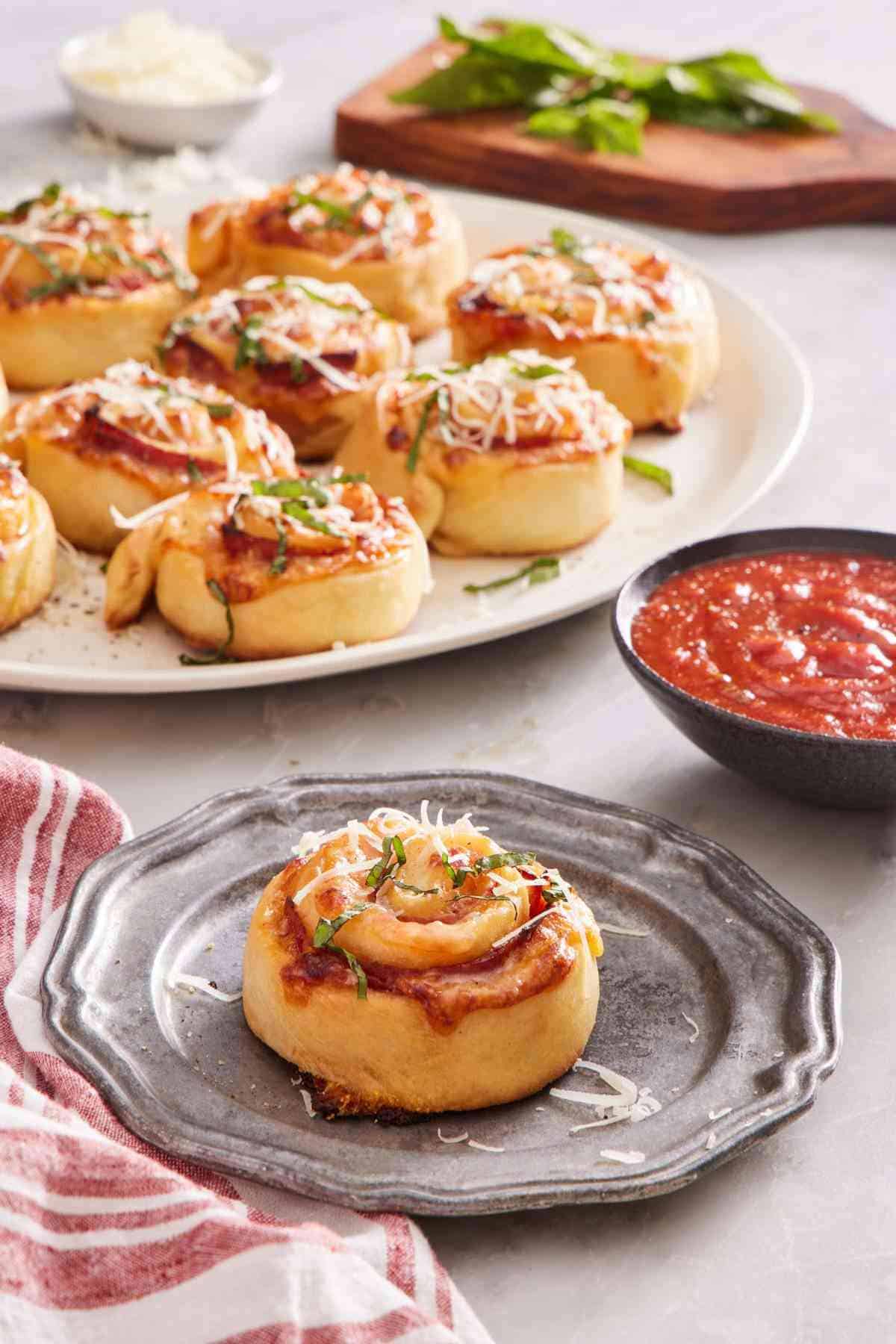 A pizza roll on a grey plate with a bowl of sauce to the side, with more homemade pizza rolls slightly out of focus in the background.