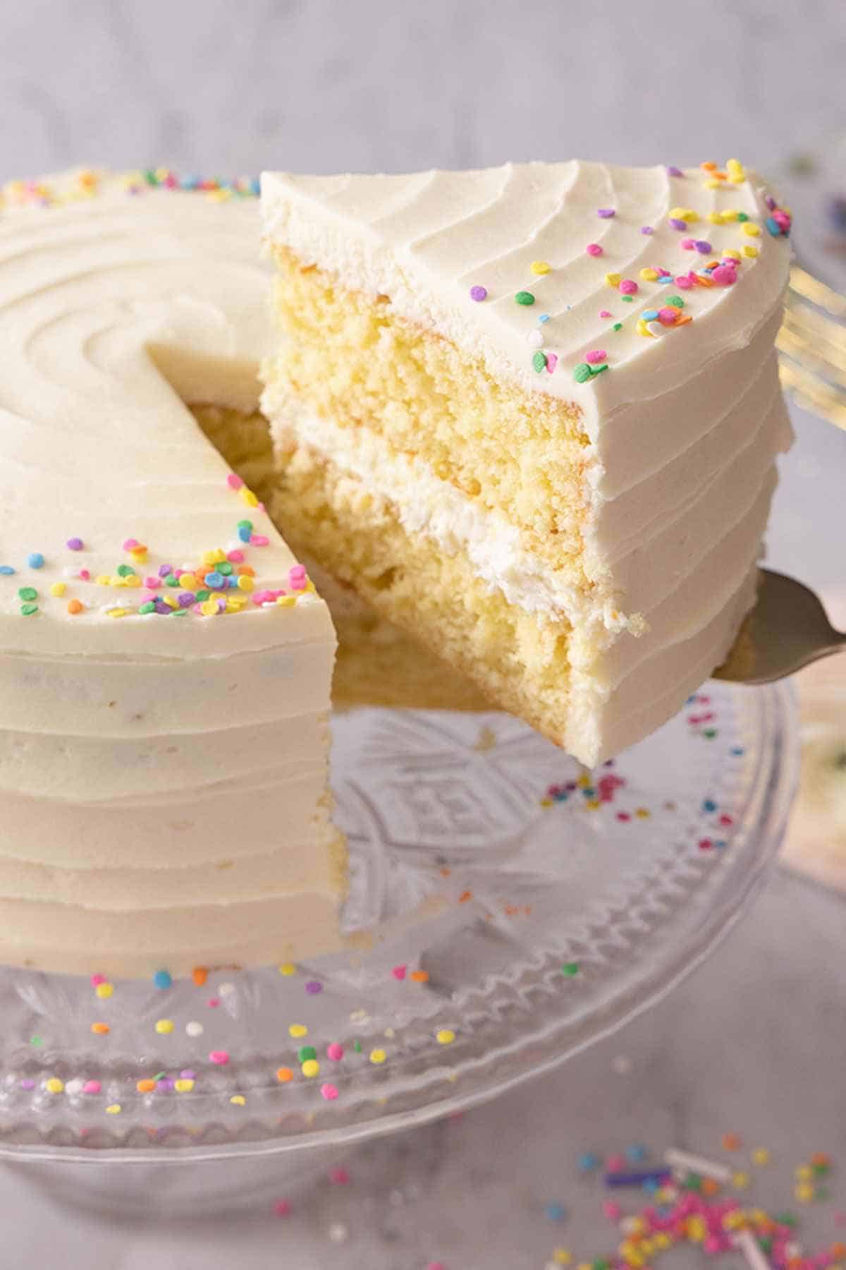 A slice of vanilla cake lifted off a cake stand holding the rest of the cake.