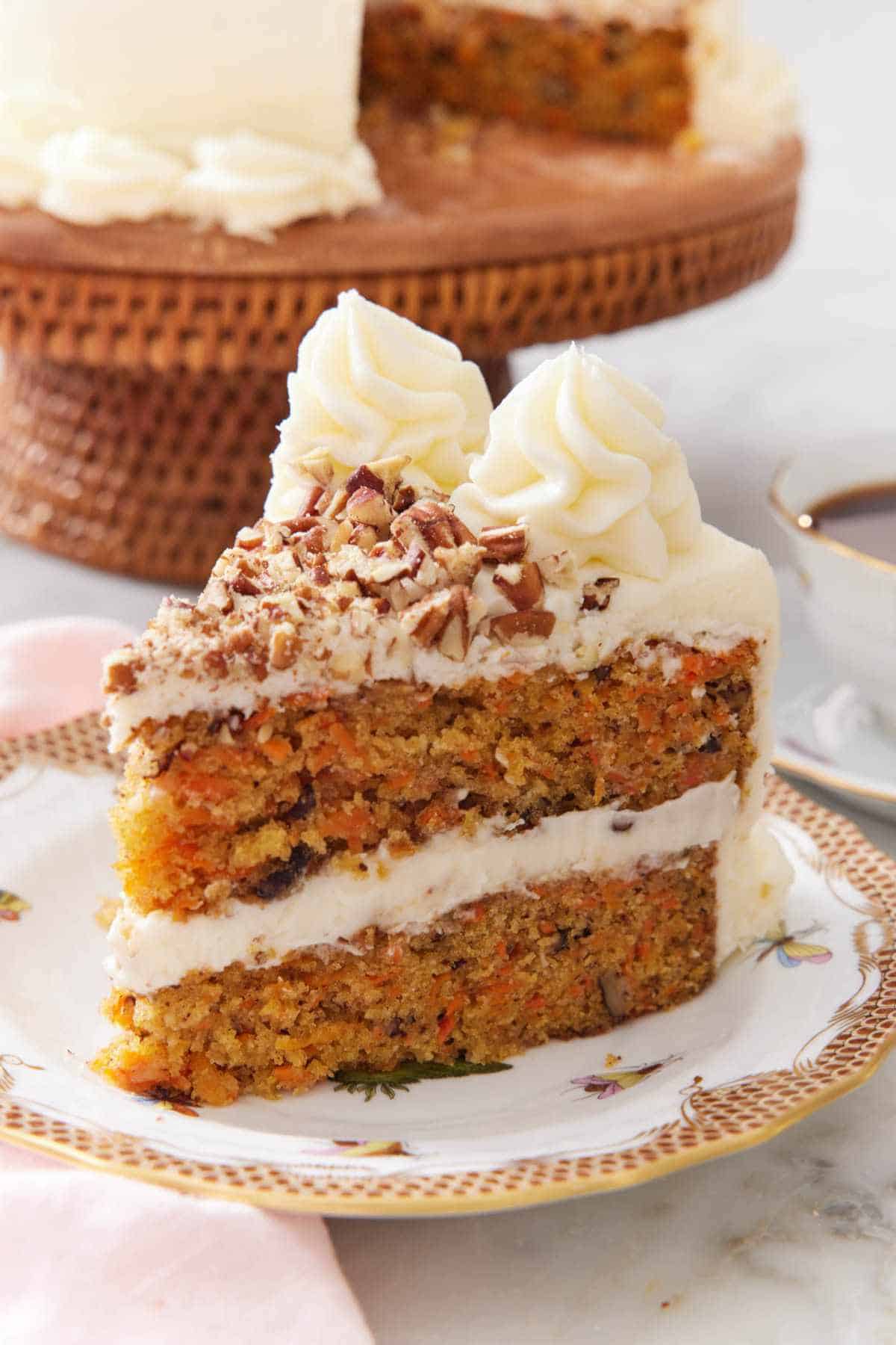 A slice of carrot cake on a small plate with the rest of the cake out of focus in the background.