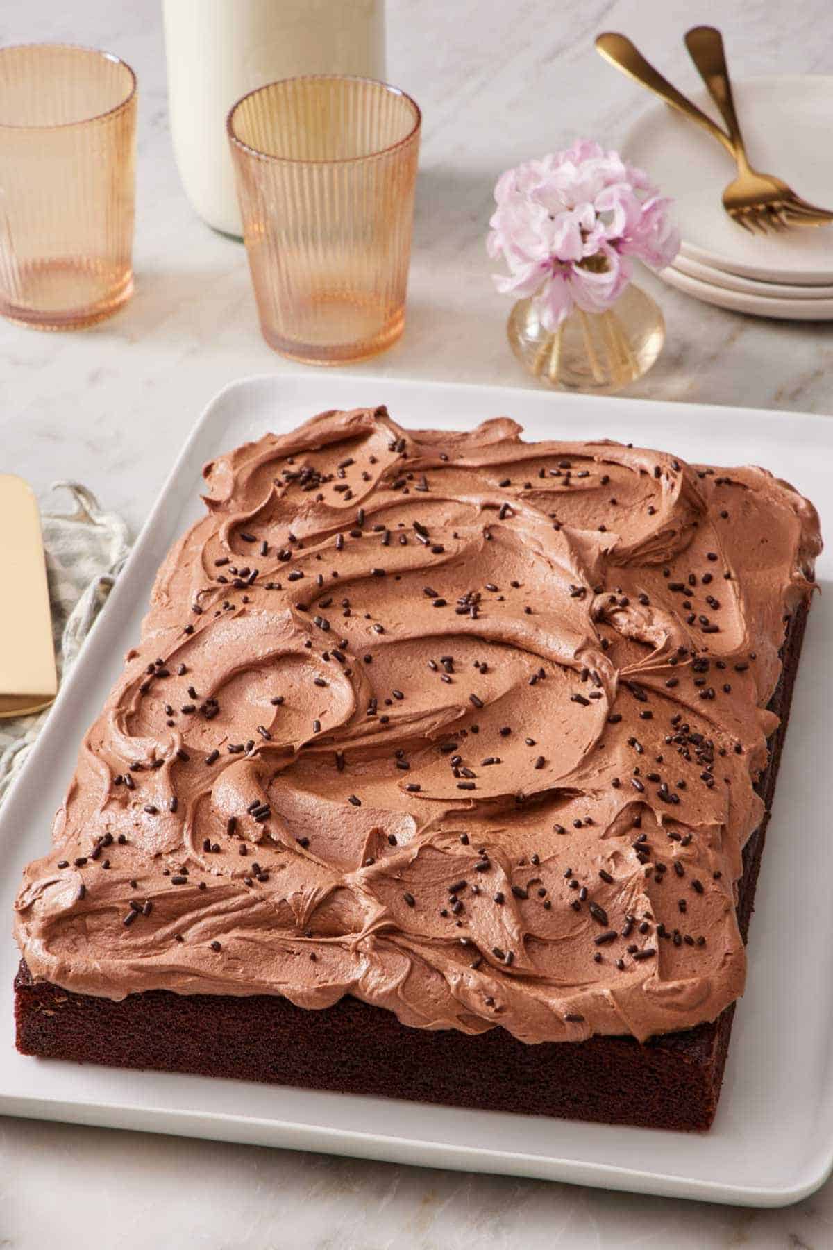 An unsliced chocolate sheet cake with frosting. Glasses and small serving plates in the background.