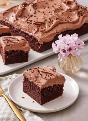 A piece of chocolate sheet cake on a small plate next to a small vase of flowers. The rest of the cake out of focus in the background.