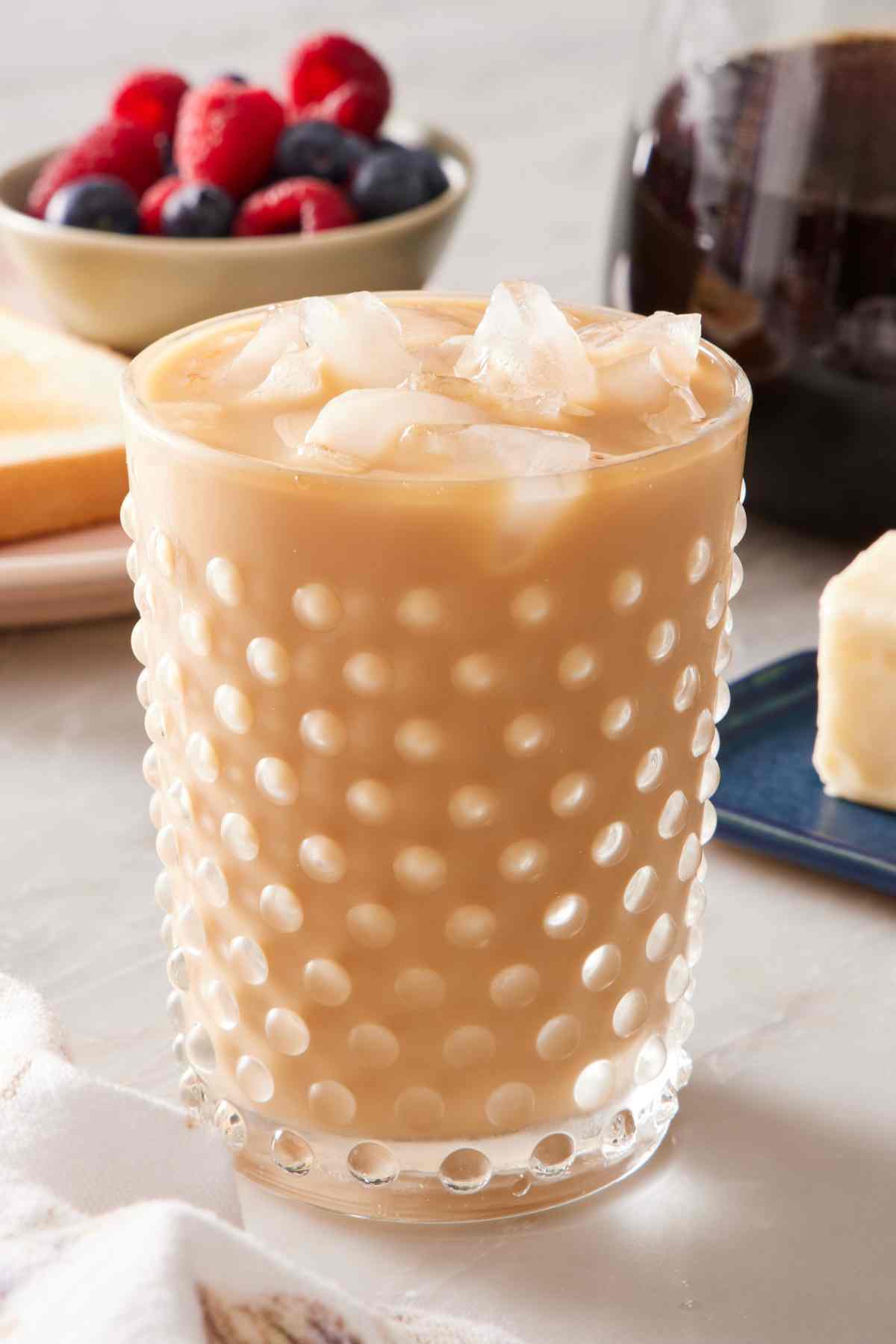 A glass of cold brew coffee with ice and milk. Berries and more coffee out of focus in the background.