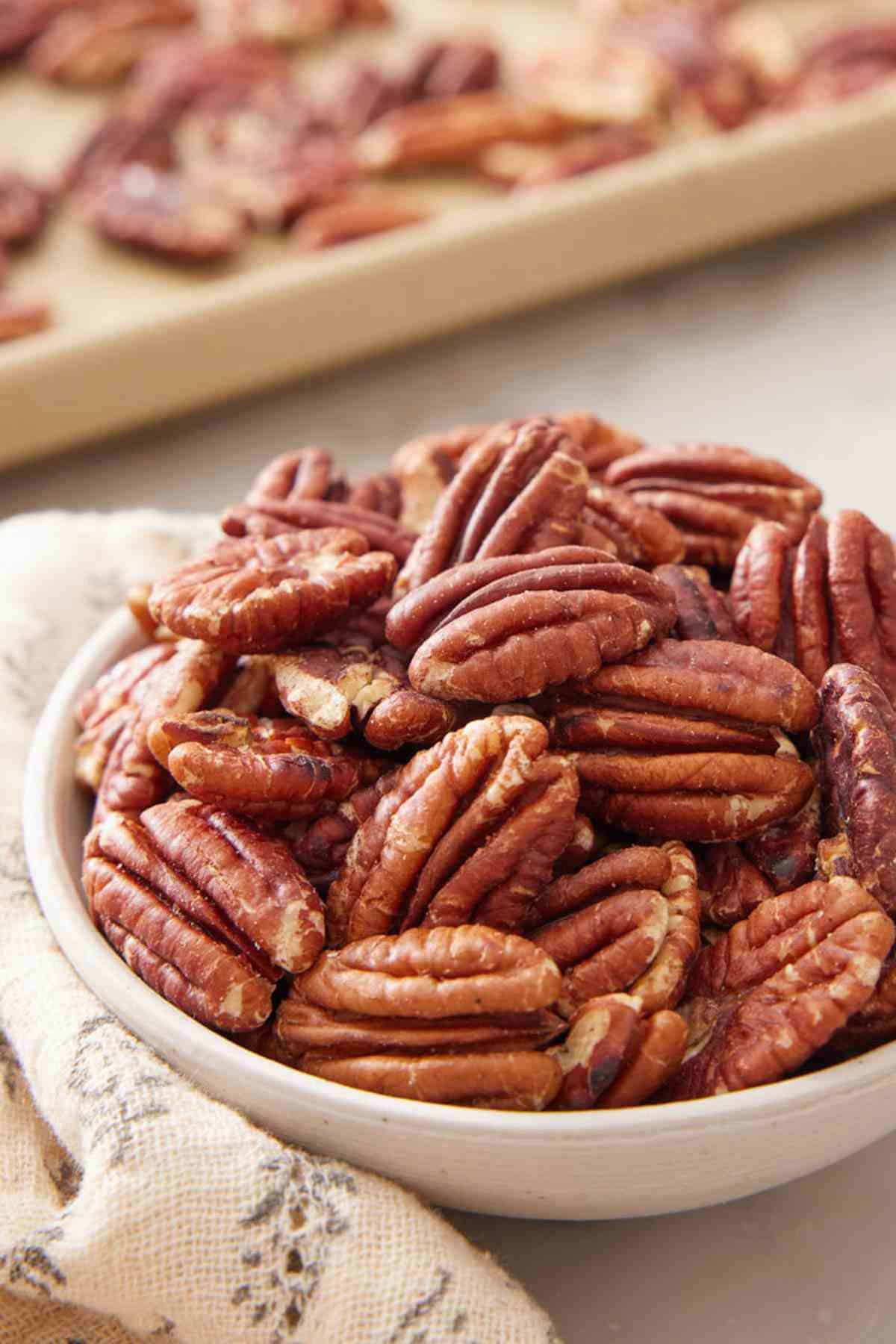 Close-up of a small bowl of toasted pecans with more on a sheet tray out of focus in the background.