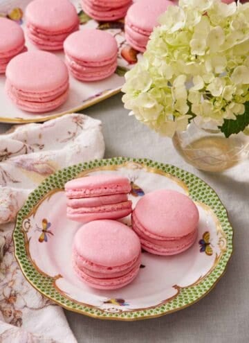 Three macarons on a plate next to a vase of flowers. A platter with more macarons in the background.