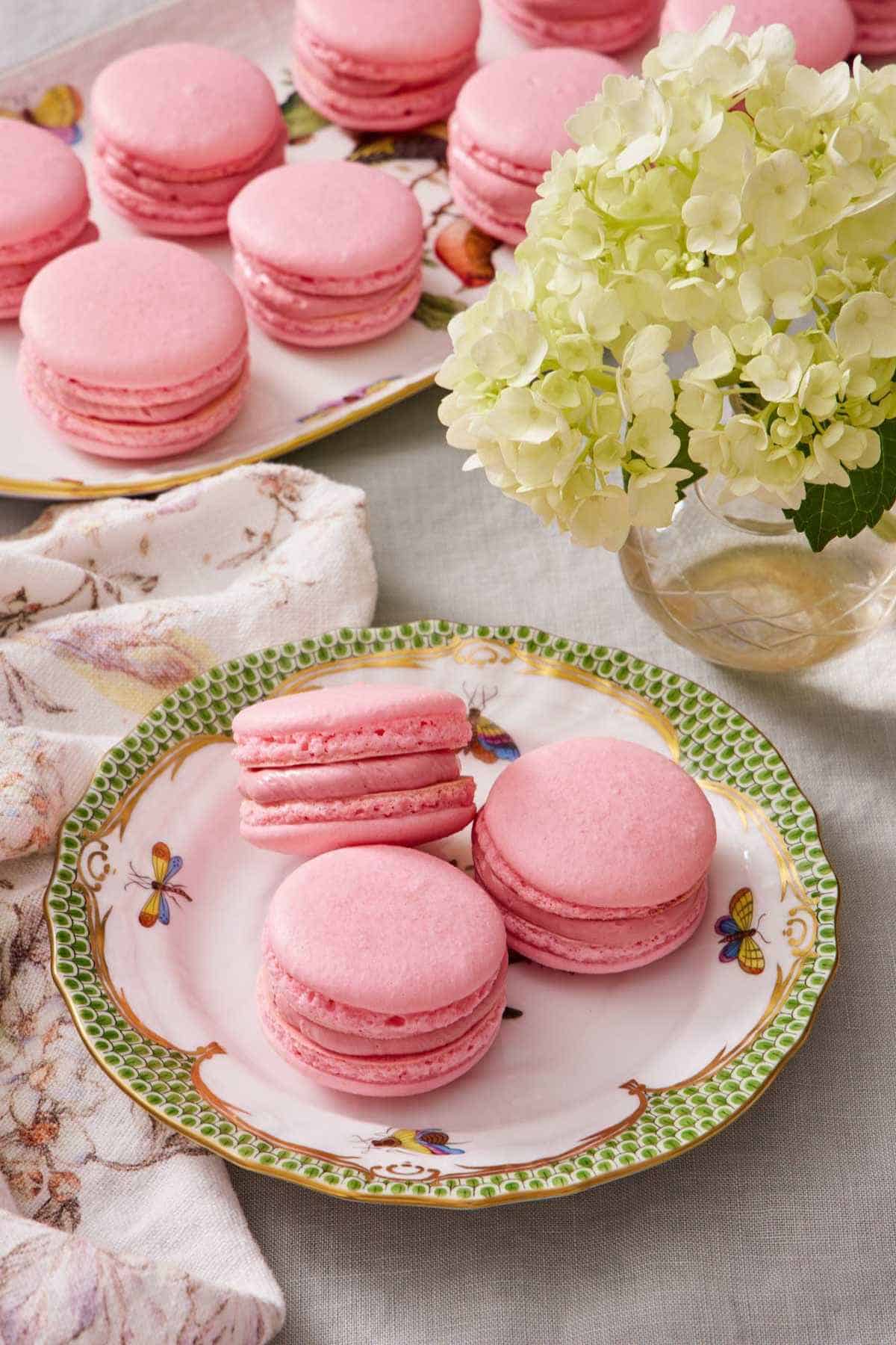 Three macarons on a plate next to a vase of flowers. A platter with more macarons in the background.