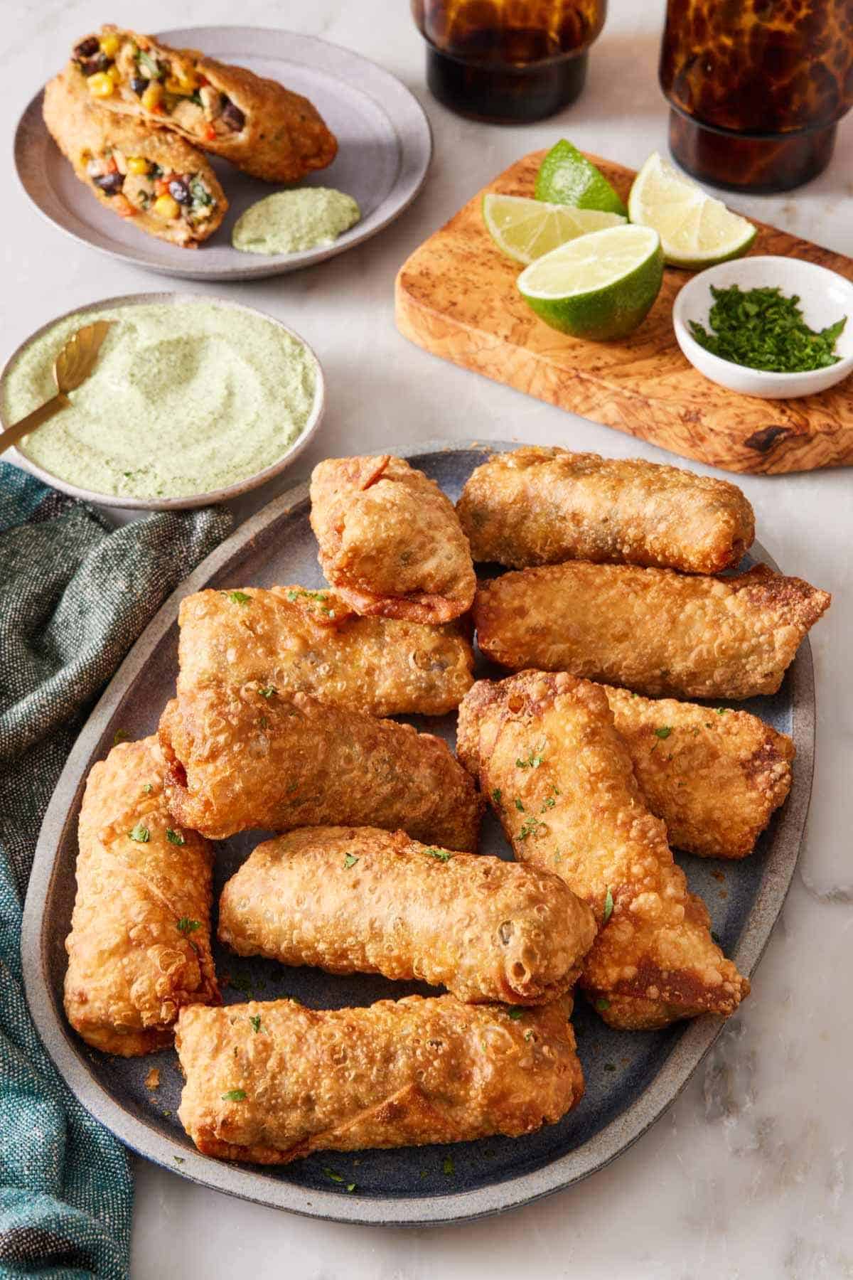 A platter of southwest egg rolls with dipping sauce, limes, and a halved egg roll behind it.