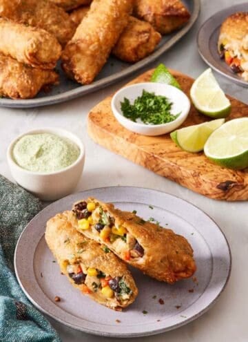A southwest egg roll on a plate, halved to showcase the filling. Limes, dipping sauce, and more egg rolls in the background.