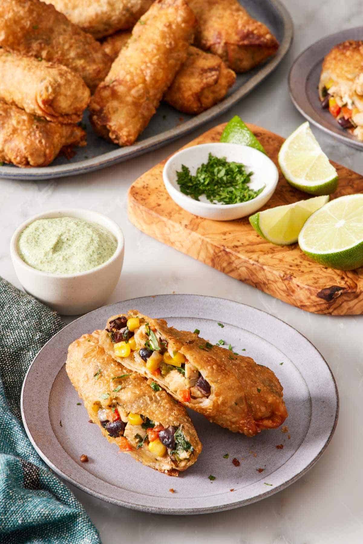 A southwest egg roll on a plate, halved to showcase the filling. Limes, dipping sauce, and more egg rolls in the background.