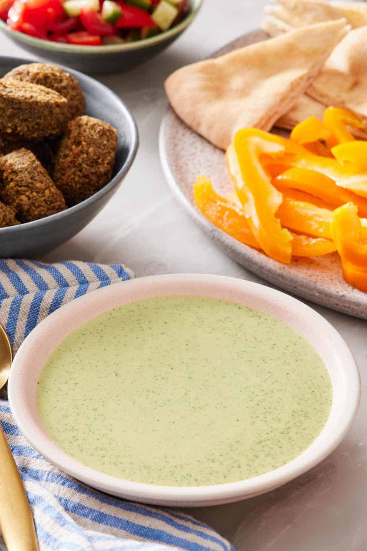 A bowl of green tahini sauce with cut vegetables, falafel, and pita in the background.
