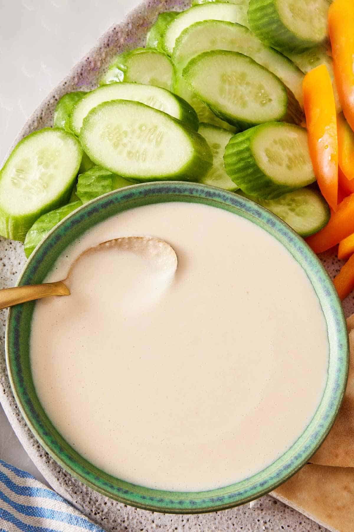 Overhead shot of a bowl of tahini sauce on a platter with cut cucumber and bell peppers.