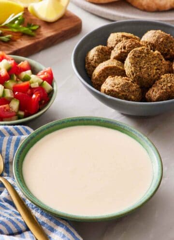 A bowl of easy tahini sauce with a bowl of falafel and another bowl of cut tomatoes and cucumber behind it.