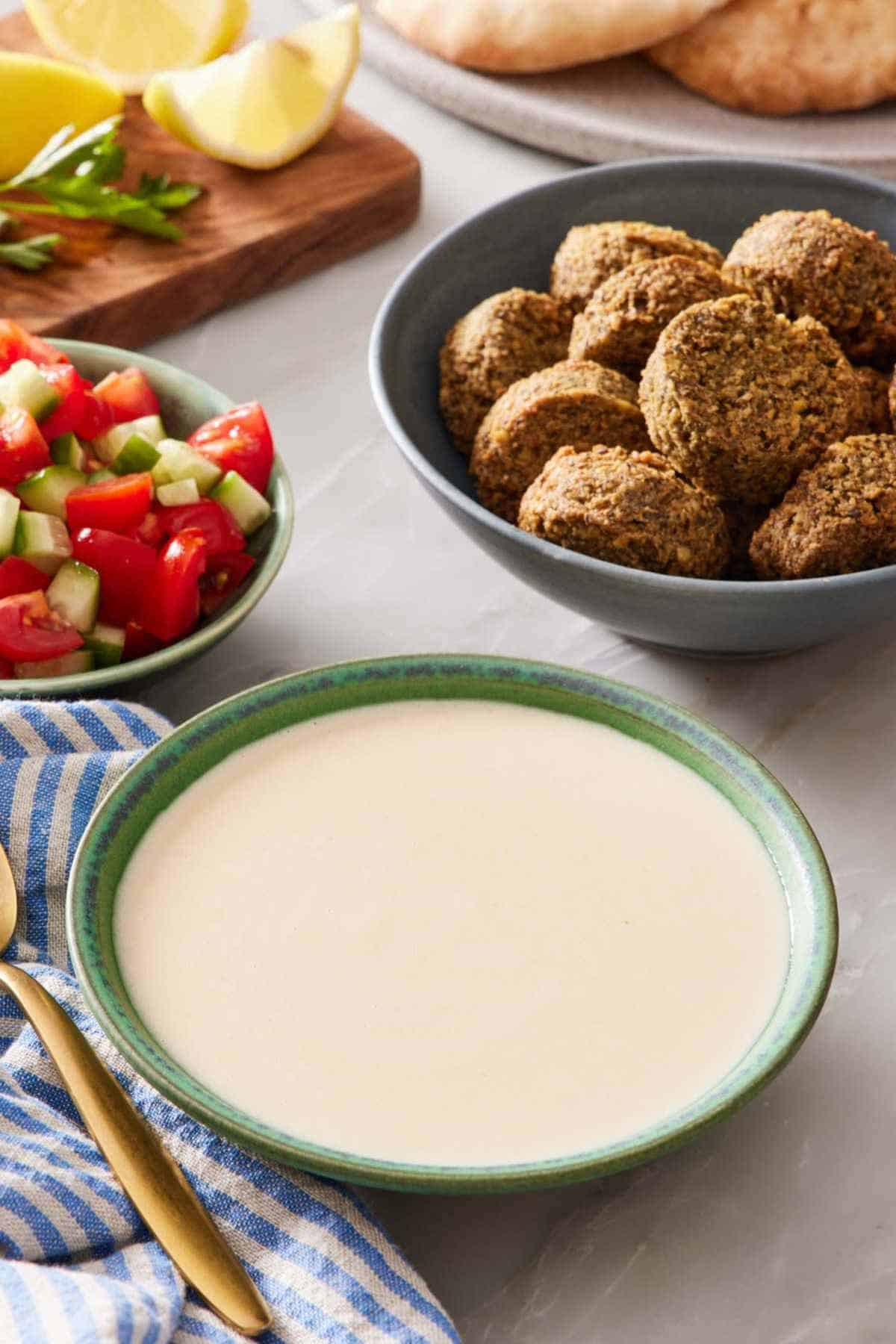 A bowl of easy tahini sauce with a bowl of falafel and another bowl of cut tomatoes and cucumber behind it.