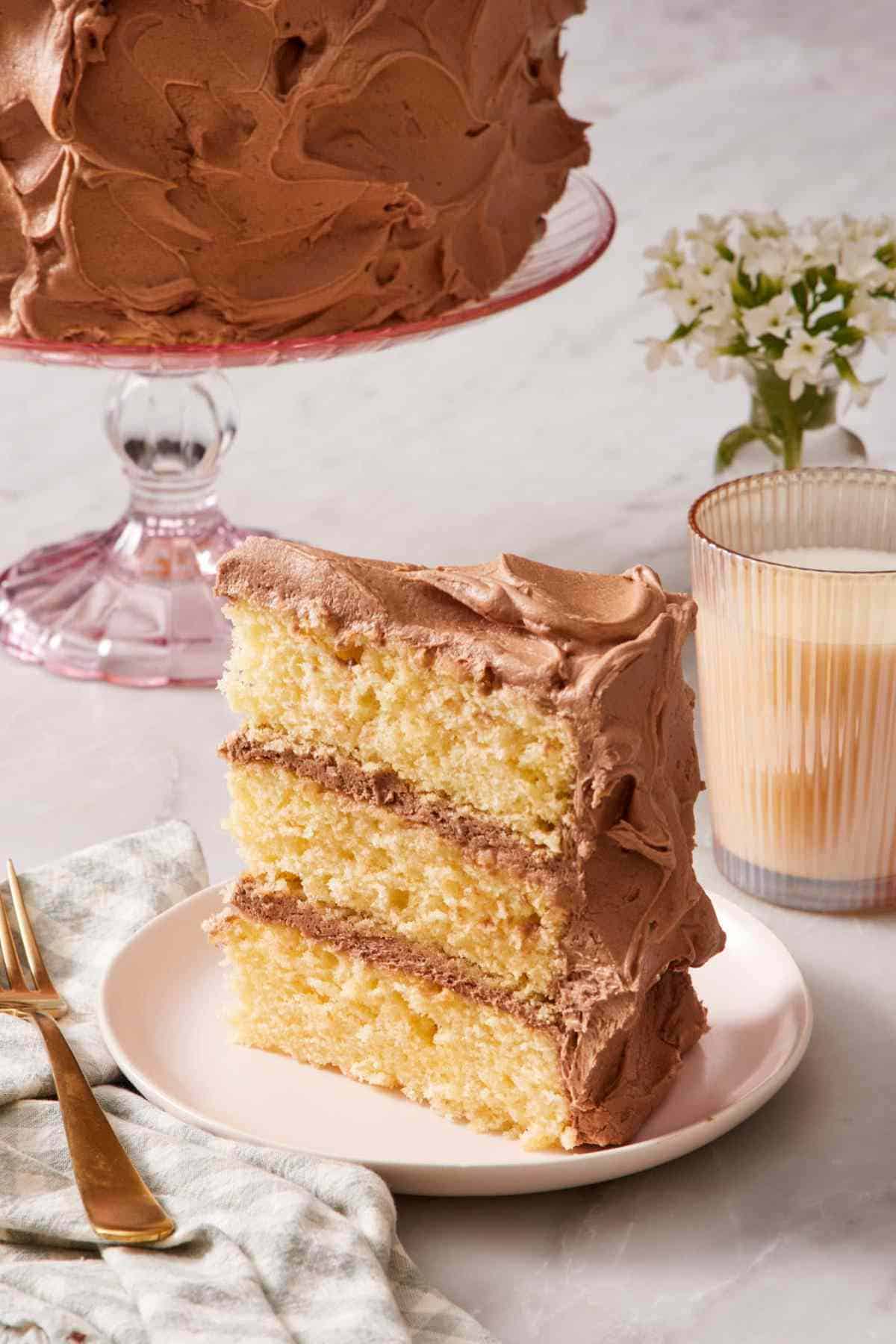 A slice of yellow cake standing up on a small plate alongside a glass of milk.