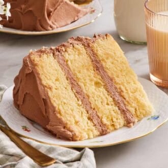 A slice of 3-layer yellow cake on its side on a plate with a glass of milk on one side, and a fork on the other.