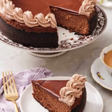 A slice of chocolate cheesecake on a small plate with the rest of the cheesecake on a cake stand right behind it.