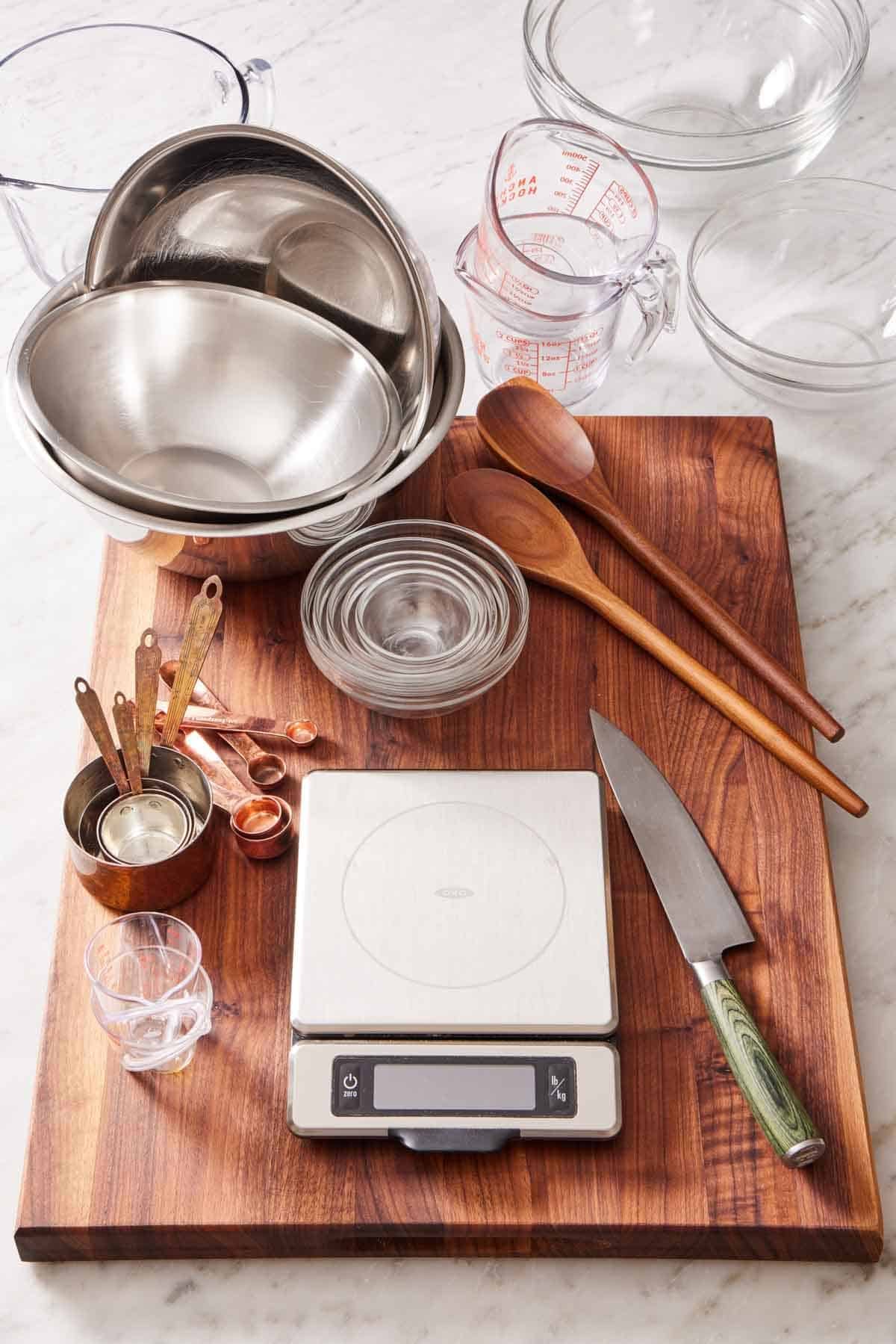 A kitchen scale, knife, measuring cups and spoons, bowls of various sizes, and wooden spoons on a wooden cutting board.