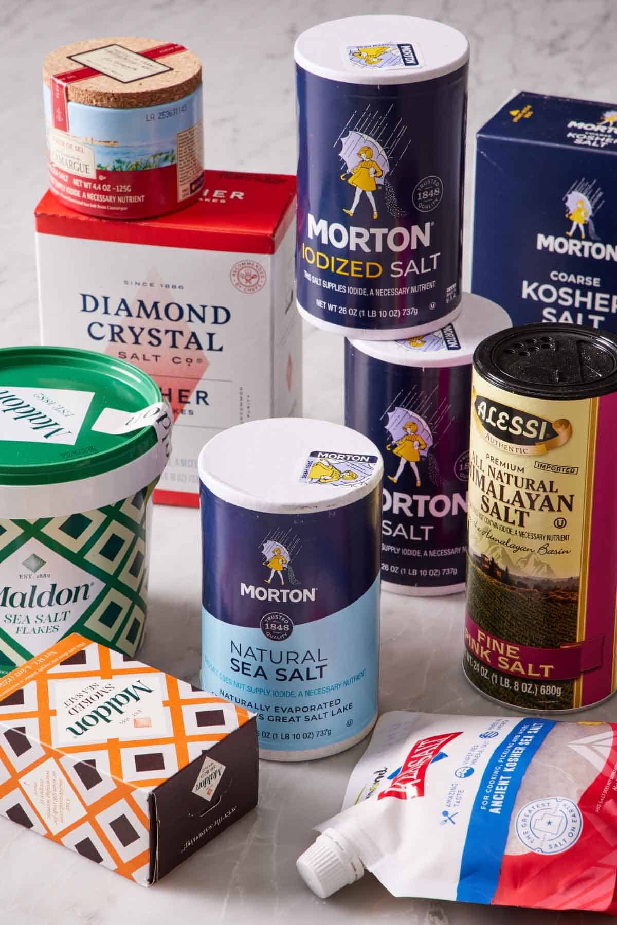 Different salt brands in their original packaging on a marble countertop.