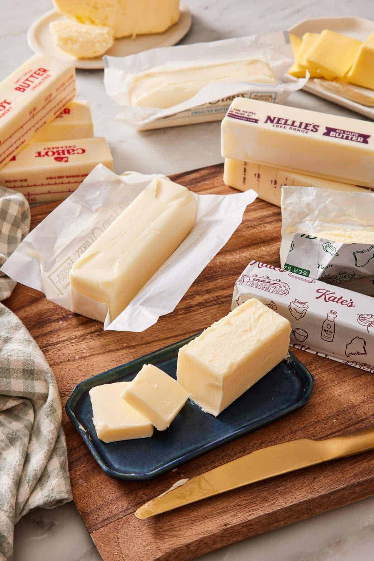 Various brands of butter sticks on a wooden cutting board and marble countertop, with one stick on a butter dish with pats cut off and stacked next to it.