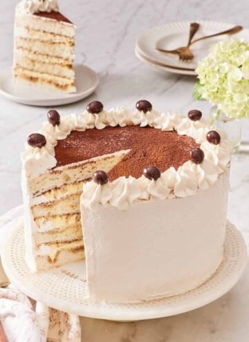 Tiramisu cake on a cake stand with a slice removed and placed on a plate in the background.