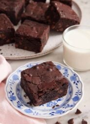 A plate with a piece of brownie with a cup of milk and platter of brownies in the background.